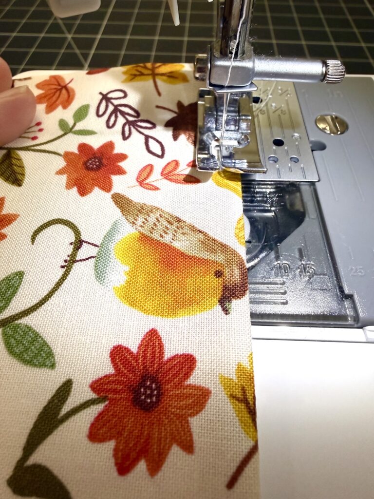 A sewing machine needle positioned over fabric patterned with autumn leaves and a bird.