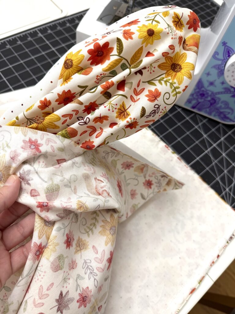 A hand holds a piece of fabric featuring an autumnal print of sunflowers, leaves, and acorns, positioned in front of a sewing machine on a cutting mat.