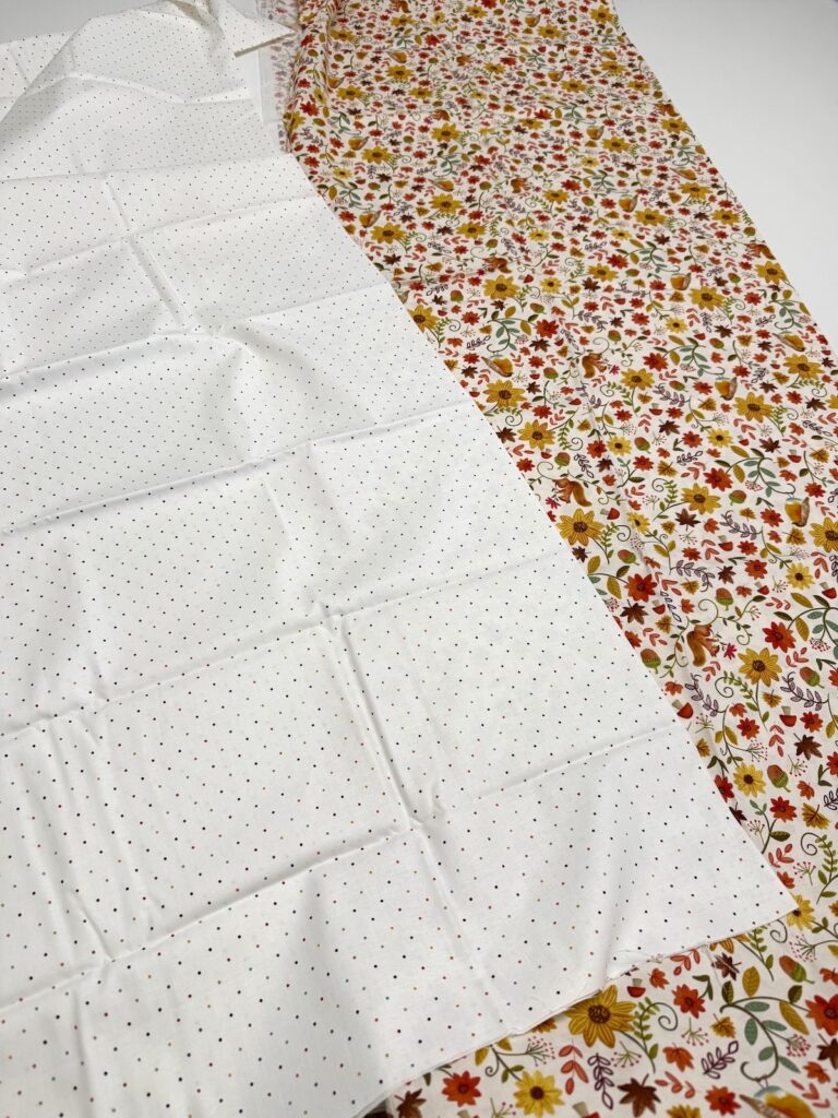 Two bolts of cotton fabric: one white with small multicolored polka dots and one with a dense floral and leaf pattern in autumn colors.